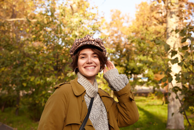 城市快乐的年轻漂亮的短发黑发女性 穿着海沟 针织毛衣和豹纹帽子 在城市花园里摆姿势 心情愉快 笑容灿烂帽子鲍勃心情