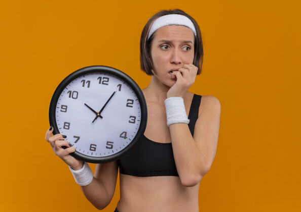 运动装身穿运动服的年轻健身女 戴着头巾 拿着挂钟 紧张地站在橙色的墙上咬指甲看人女人