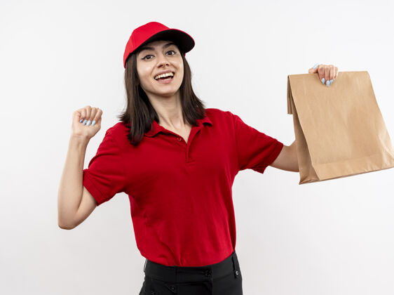 举行年轻的送货员身穿红色制服 戴着帽子 手里拿着纸包 握紧拳头 站在白色的背景上 开心而兴奋握紧兴奋帽子