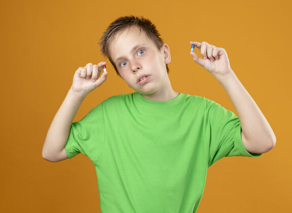 男孩生病的小男孩穿着绿色t恤 身体不适 手里拿着药片 站在橙色的背景上 看着摄像机 很困惑不适小T恤