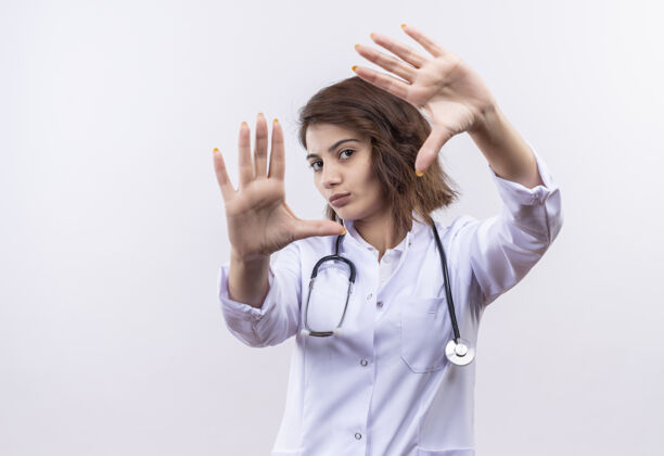 手穿着白大褂的年轻女医生用听诊器做了一个框架 双手穿过这个框架 严肃的脸站在白色的墙上长相做外套