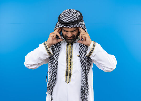 站一位身着传统服装的阿拉伯男子站在蓝色的墙上 摸着太阳穴 看上去很不舒服 头痛得厉害触摸头痛强壮