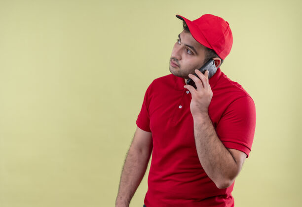 帽子年轻的送货员穿着红色制服 戴着帽子 站在绿色的墙上 一边看手机一边严肃地看着严肃送货说话