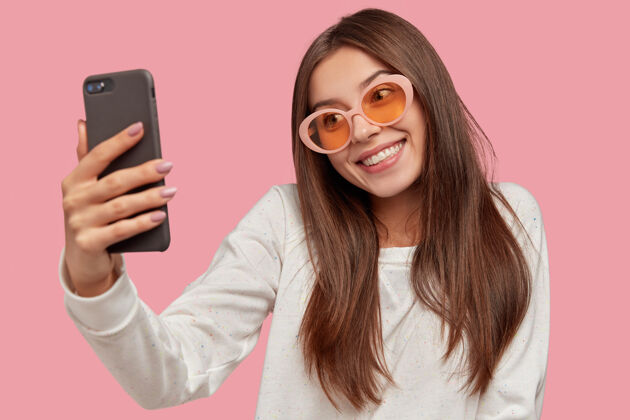 年轻横拍的漂亮黑发女孩开心地笑着 歪着头 拿着手机 录视频 自拍 戴着时髦的太阳镜和白色休闲毛衣 隔着粉色的墙采取手机肖像