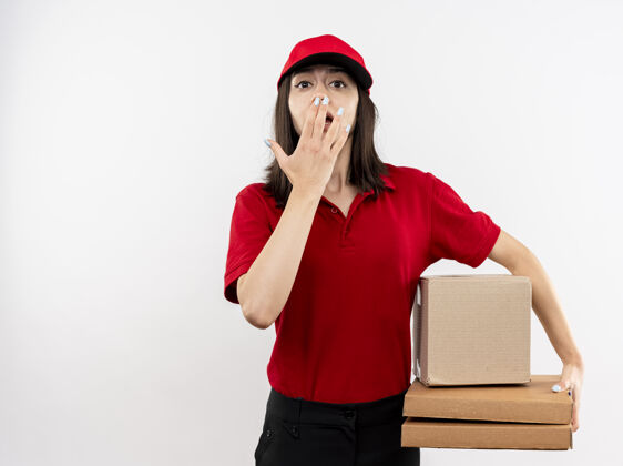 站着年轻的送货女孩穿着红色制服 戴着帽子 拿着盒子和披萨盒 看着镜头 惊讶地捂着嘴 手站在白色的背景上嘴盖年轻
