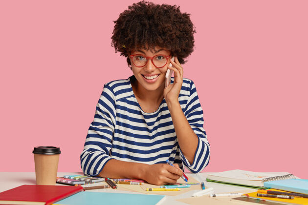 智能手机成功的黑皮肤女人 留着非洲式发型 穿着条纹衣服 一边用笔记本画东西 一边打电话 一边喝咖啡 感觉很满足 很有灵感项目室内记事本
