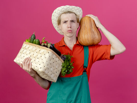 蔬菜年轻的园丁穿着连体衣戴着帽子拿着装满蔬菜的箱子穿连身衣脸