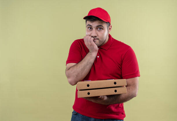 盒子年轻的送货员身穿红色制服 头戴鸭舌帽 手里拿着披萨盒 紧张地咬着指甲站在绿色的墙上男人人人