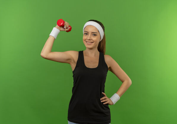 人身穿运动服 头箍 举着哑铃的年轻健身女士站在绿色的墙上 脸上带着微笑 看起来很自信头带运动装站着