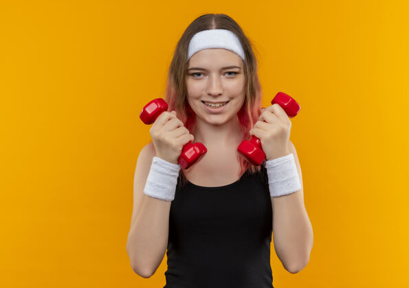 健身身着运动服的年轻健身女士举着哑铃 微笑着站在橙色的墙上做运动运动装运动员市民