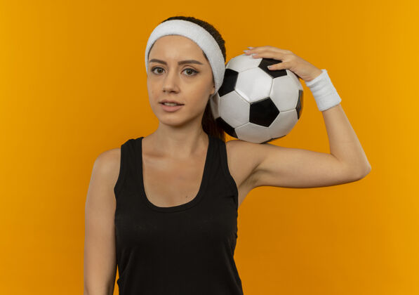 运动身穿运动服 头箍 手持足球的年轻健身女士站在橙色的墙上 看起来很自信公动装站着