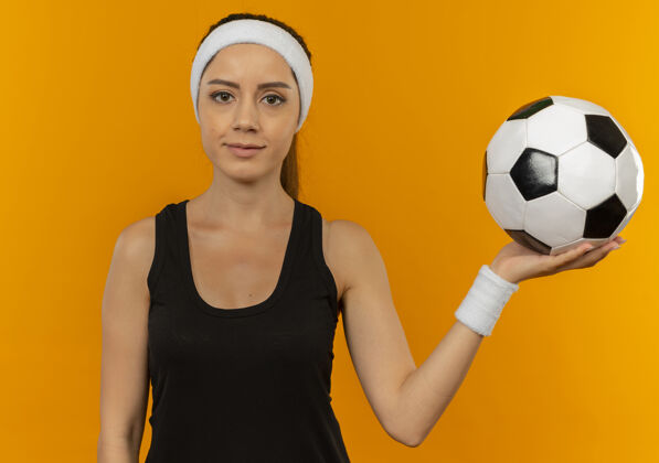 年轻身穿运动服 头戴钢带 正经脸举着足球站在橙色墙壁上的年轻健身女士运动员抱着运动装