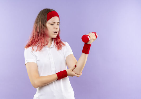 感觉年轻的运动女子举着一个哑铃做运动看着手肘感觉疼痛站在紫色的墙上年轻人看