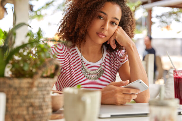 信息严肃的黑人女性形象在手机上阅读收到的信息 在手机上查看电子邮件 拿着咖啡杯坐在咖啡馆内部 连接无线互联网 购物手机咖啡馆认真