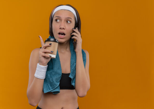 年轻身穿运动服 脖子上套着毛巾 手里拿着咖啡杯 站在橘色墙上讲手机的年轻健身女士 看上去很困惑拿着咖啡脖子