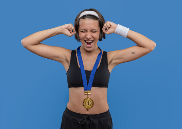 成功一位身穿运动服 脖子上戴着金牌的年轻女子握紧拳头 为自己站在蓝色墙壁上的成功而欢欣鼓舞运动员高兴人