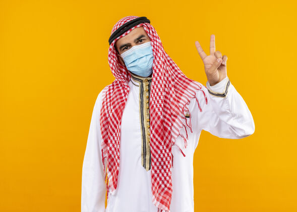 阿拉伯语身着传统服装和面罩的阿拉伯商人 自信的表情 站在橙色的墙上展示胜利的标志穿着传统商务人士
