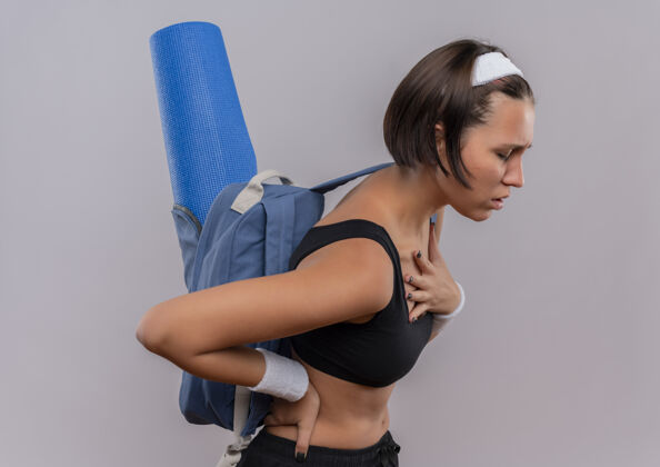手穿着运动服 背着背包和瑜伽垫 手放在胸口看起来不舒服的年轻健身女士站在白墙上累了人市动员