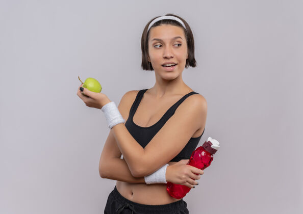 健康身穿运动服的年轻健身女士手持青苹果和一瓶水 自信地微笑着站在白色的墙上望向一边女人站着人