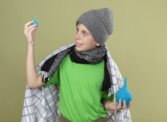 微笑生病的小男孩戴着暖和的帽子 围着围巾 裹着毯子 拿着灌肠剂 站在明亮的背景下微笑着看着他们帽子小抱着