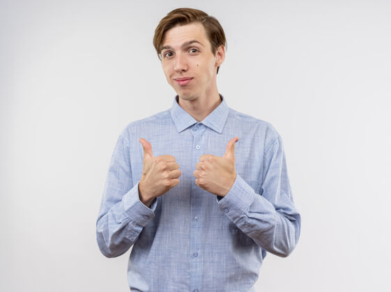 脸身穿蓝色衬衫的年轻人站在白色背景上 面带微笑地竖起大拇指看着摄像机年轻拇指微笑