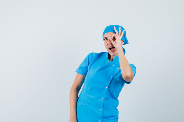 年轻的身穿蓝色制服的女医生 眼睛上显示ok标志 看上去很惊讶 正视图治疗女人成人
