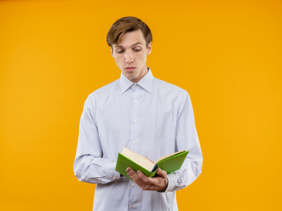 阅读一个穿着白衬衫的年轻人拿着一本打开的书 站在橙色的背景上 带着怀疑的表情看着书握着年轻站着