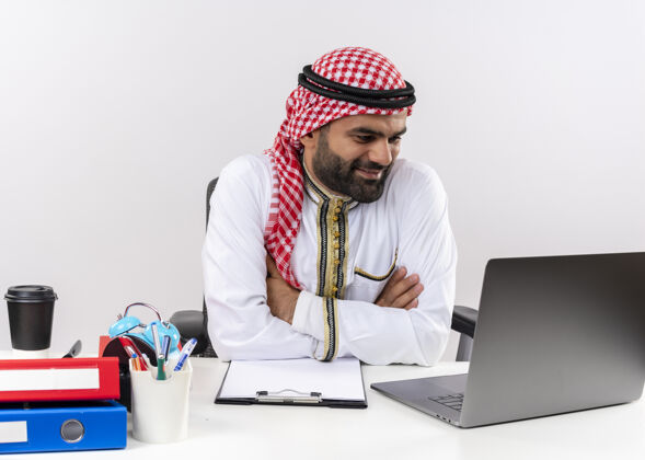 电脑身着传统服装的阿拉伯商人坐在桌旁 拿着笔记本电脑看着屏幕微笑着在办公室工作看传统工作