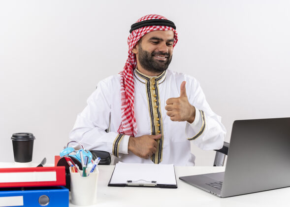 拇指身着传统服装的阿拉伯商人坐在桌旁 手持笔记本电脑 开心而积极地微笑着 竖起大拇指在办公室工作桌子展示商务人士