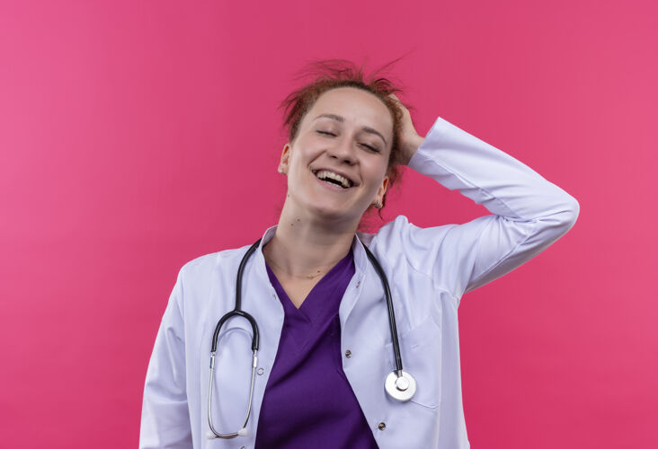 年轻人年轻的女医生身穿白大褂 手持听诊器 微笑着 手放在头上 站在粉红色的墙上 快乐而高兴头听诊器医生