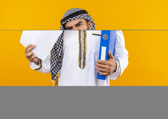 空白阿拉伯商人在传统穿拿文件夹和空白页不高兴站在橙色的墙上看穿商务人士