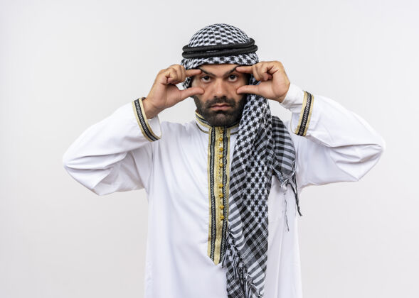 站传统的阿拉伯男人戴着张开的眼睛用手指试图看得更好站在白墙上试试手指传统