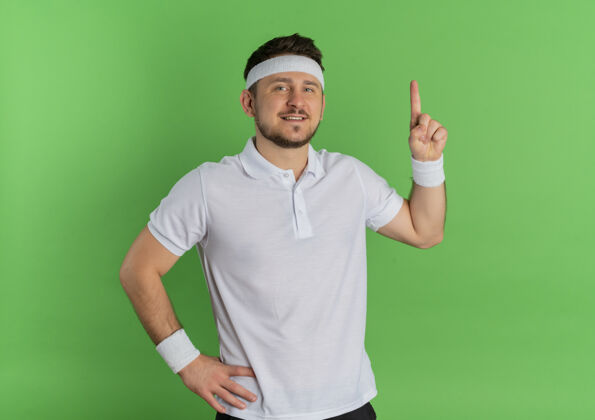 人身穿白衬衫 头戴头巾的年轻健身男子面带微笑 自信地指着绿色的墙壁运动员年轻运动装