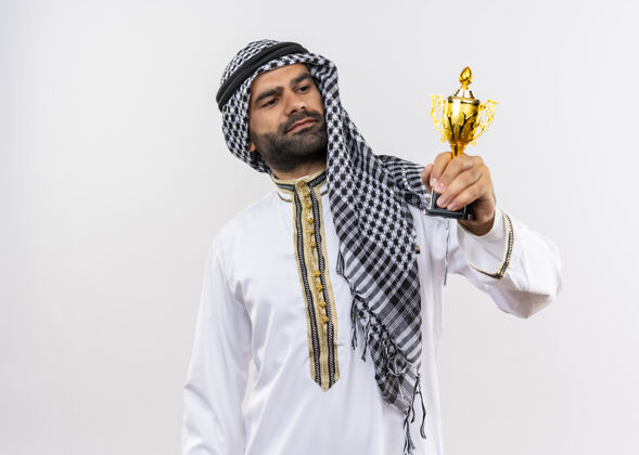 看身着传统服饰的阿拉伯男子手持奖杯 高傲地站在白色的墙上看着奖杯举行男子传统