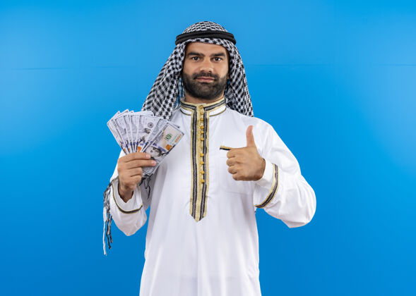 传统身着传统服装的阿拉伯男子站在蓝色的墙上 露出现金微笑 自信地竖起大拇指自信显示现金
