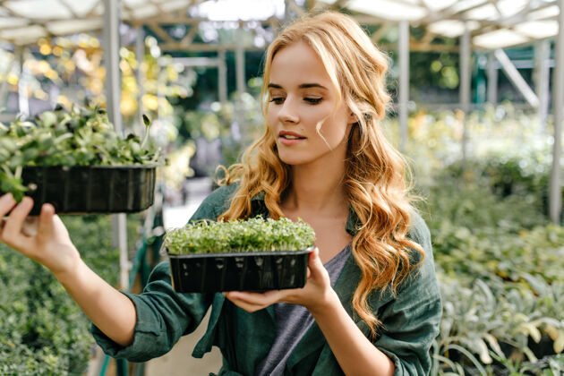 金发女孩在外面拿着两个装有绿色小植物的塑料罐的画像年轻的女植物学家研究绿色植物积极成人明亮