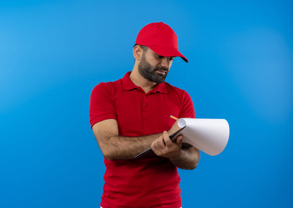 站着留着胡子的送货员穿着红色制服 戴着帽子 手里拿着剪贴板 手里拿着空白页 严肃地站在蓝色的墙上看着他们脸胡子制服