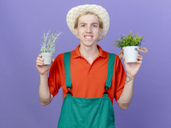 罐子高兴的年轻园丁男子穿着连体衣和帽子举行盆栽植物连身衣年轻人抱着