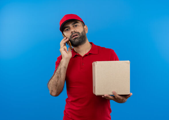 制服留着胡子的送货员穿着红色制服 戴着帽子拿着盒子 站在蓝色的墙上严肃地讲着手机送货站着严肃