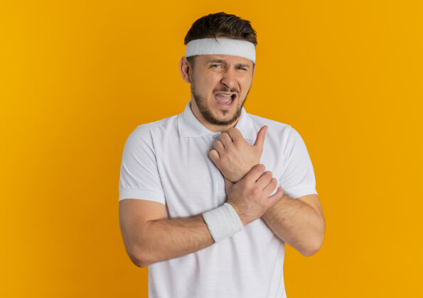 健康身穿白衬衫 头箍朝前看的年轻健身男子站在橘色的墙上 手上触碰着疼痛的手腕手腕运动头带