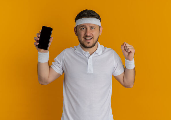 运动身穿白衬衫 头戴智能手机 握紧拳头的年轻健身男子站在橙色的墙上 快乐而积极教练站着健身