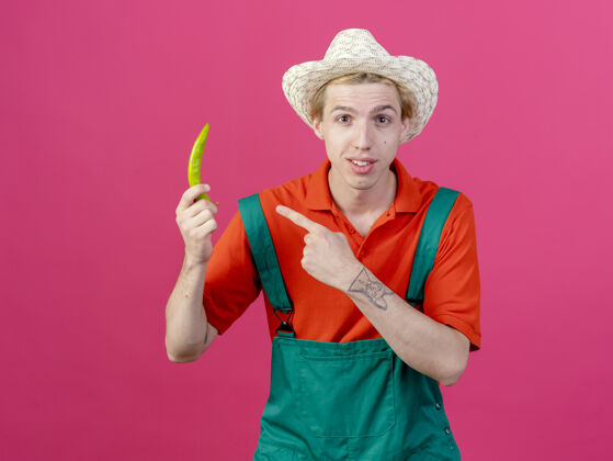 胡椒年轻的园丁 穿着连身衣 戴着帽子 指着青椒站立粉色年轻