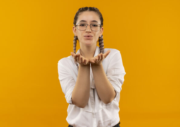 人一个戴着眼镜 梳着辫子 穿着白衬衫的年轻女学生手拉手 站在橙色的墙前 吹着吻女孩站着衬衫