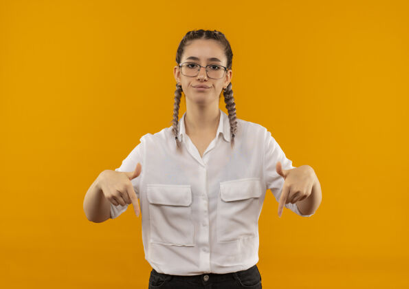 学生戴着眼镜 梳着辫子 穿着白衬衫的年轻女学生望着前面 带着怀疑的表情 手指朝下 站在橙色的墙上表情站指向