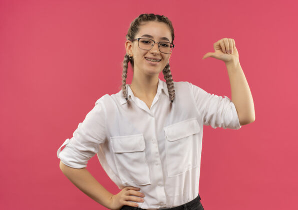 学生戴着眼镜 梳着辫子 穿着白衬衫的年轻女学生望着前方 微笑着 快乐而积极地指着自己站在粉红色的墙上积极年轻衬衫