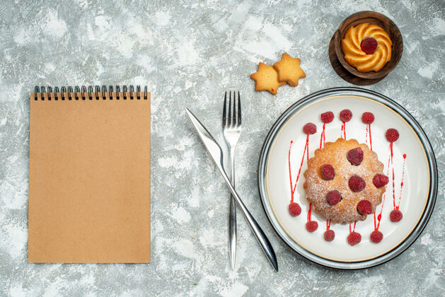 针俯瞰白色椭圆形盘子上的浆果蛋糕 碗叉上的饼干 灰色表面上的餐刀笔记本钳子晚餐手工工具