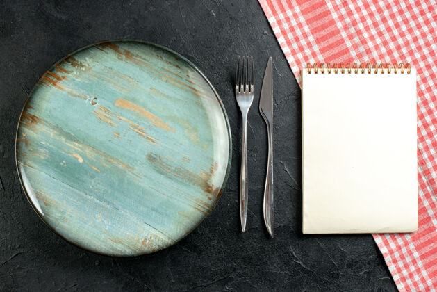 视图顶视图圆形餐叉和餐刀红白格子桌布笔记本放在黑桌子上圆盘子桌布刀