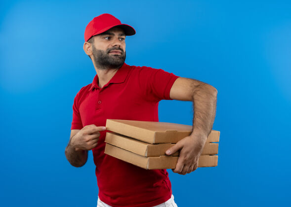 送货一个留着胡须的送货员 穿着红色制服 戴着帽子 手里拿着一叠比萨饼盒 一边看着一边 用手指着蓝色墙上的盒子看男人帽子