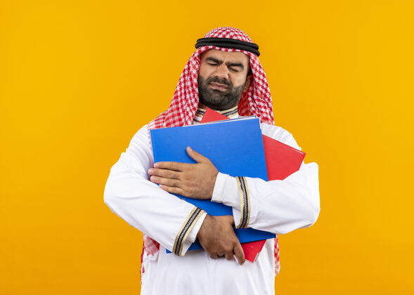 持有身着传统服装的阿拉伯商人站在橙色的墙上 手里拿着两个文件夹 看上去既不高兴又不高兴文件夹传统穿