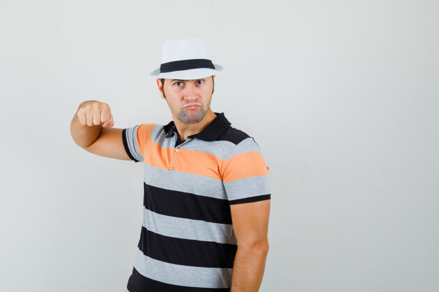 愤怒年轻人举起他的拳头在条纹t恤衫 帽子和愤怒的表情 前视图聪明男性拳头